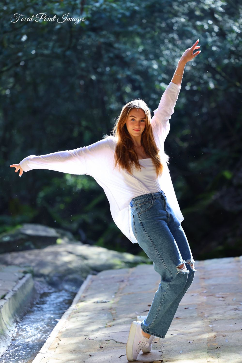 Dancer in white sweater and jeans strikes a graceful pose on rocks by a stream.
