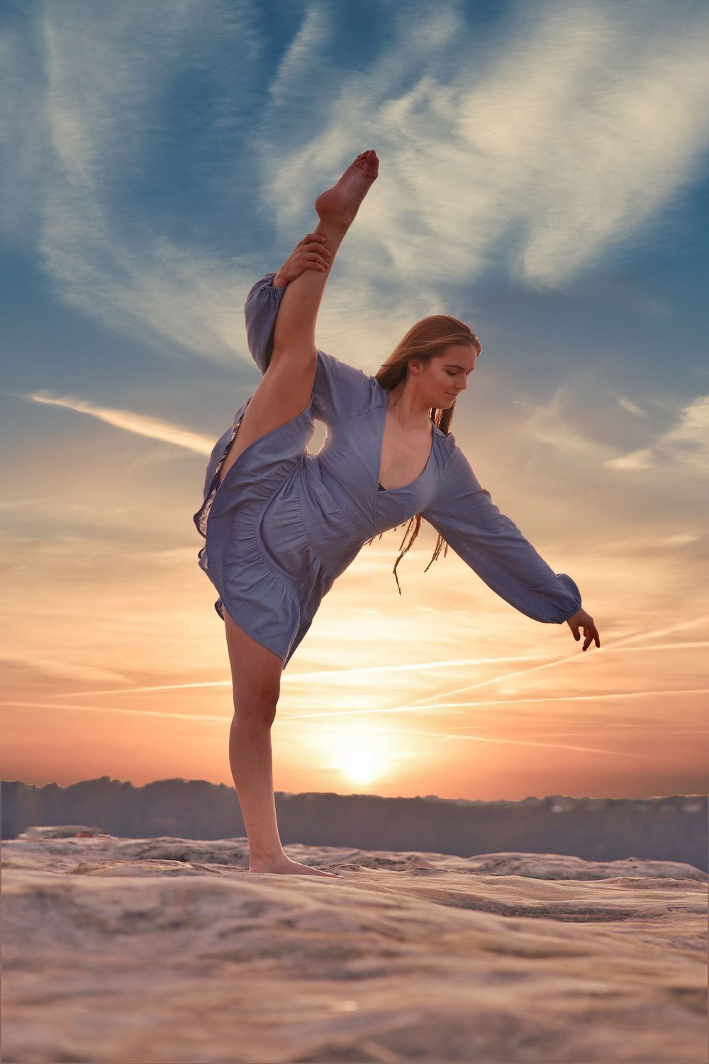 Dancer performs high kick on beach at sunset with dramatic sky backdrop.