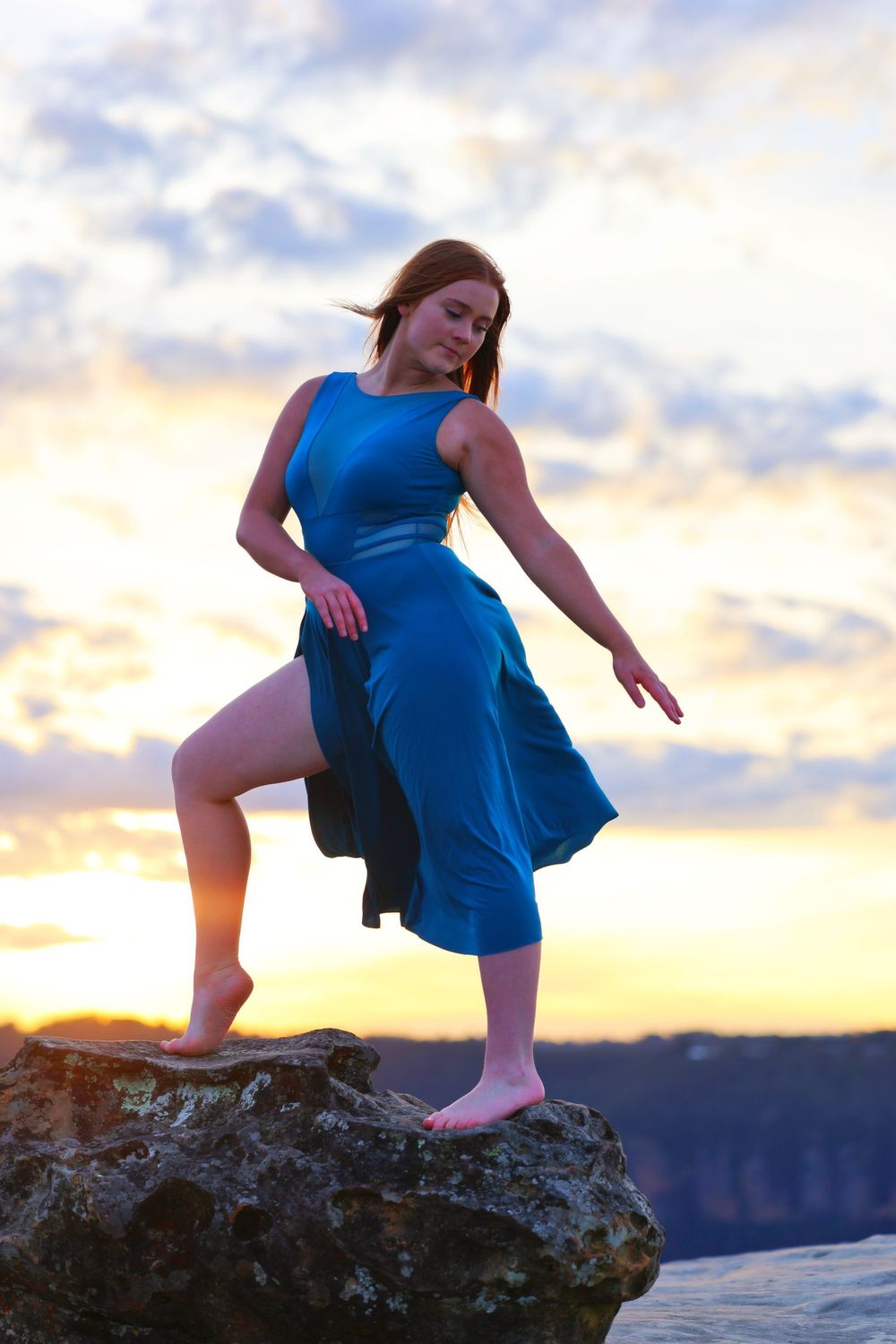 Dancer in blue dress poses dramatically on rocky cliff at sunset.