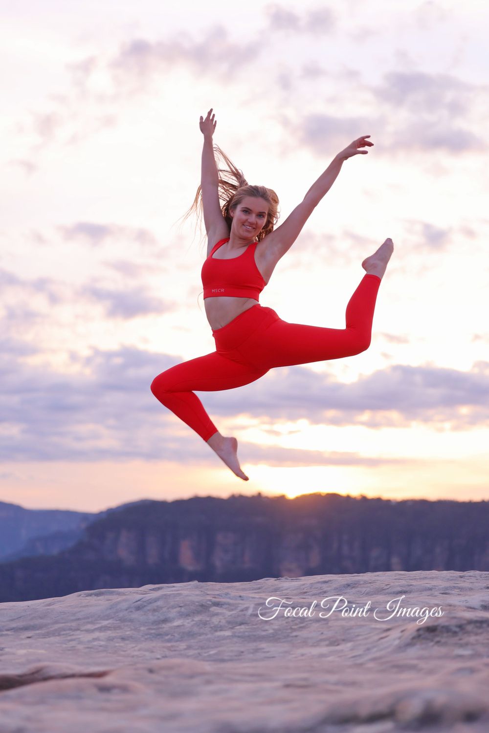 Dancer in red athletic wear performs dynamic leap against sunset mountain backdrop.
