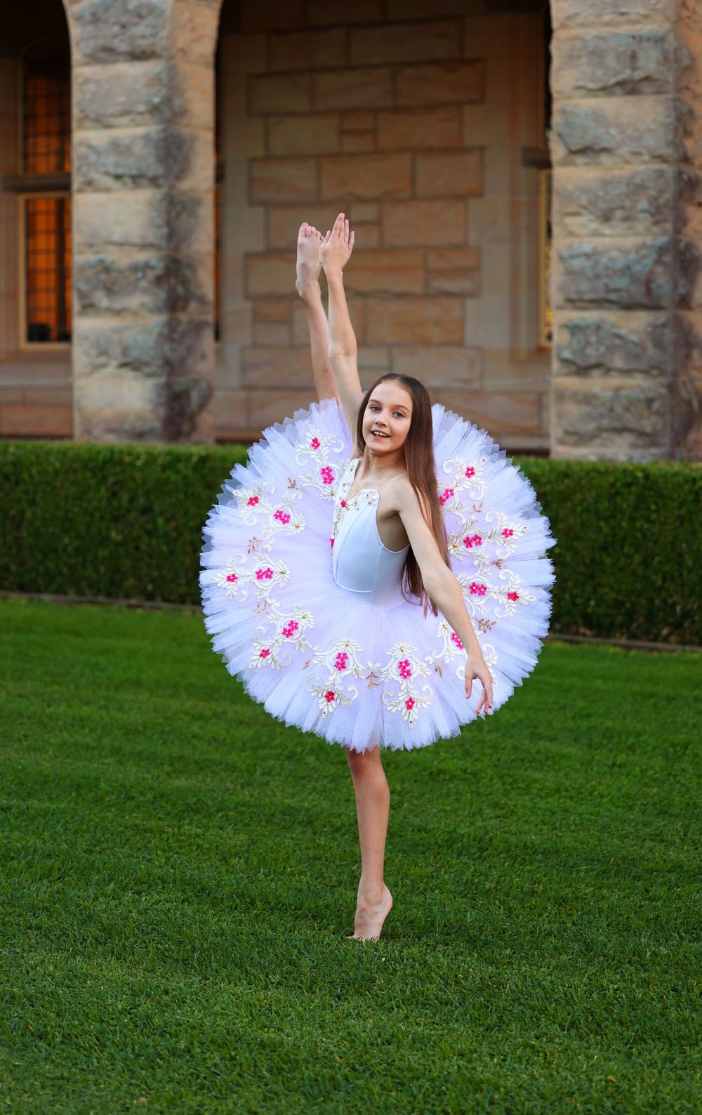Classical dancer poses in white tutu with pink accents on grassy lawn.
