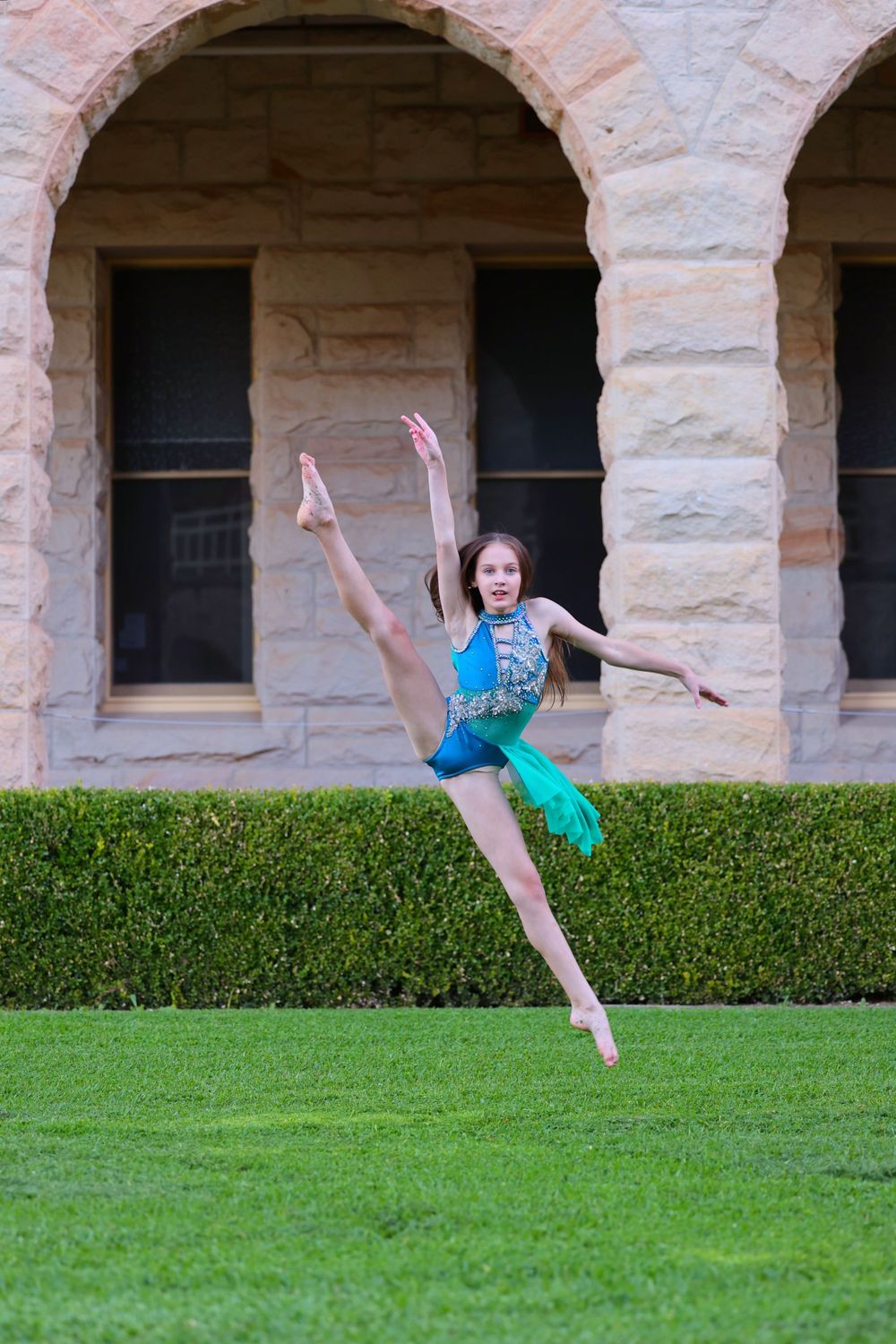 Dancer in turquoise leotard leaps gracefully in front of stone archway architecture.