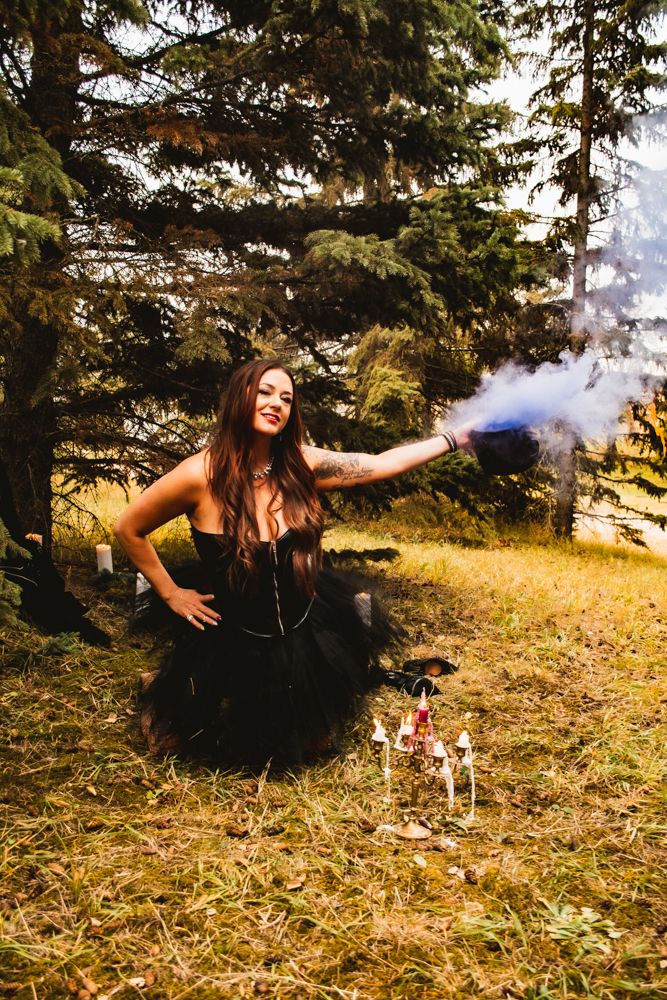 Dark tulle dress outdoor portrait with blue smoke and decorative elements among evergreen trees.
