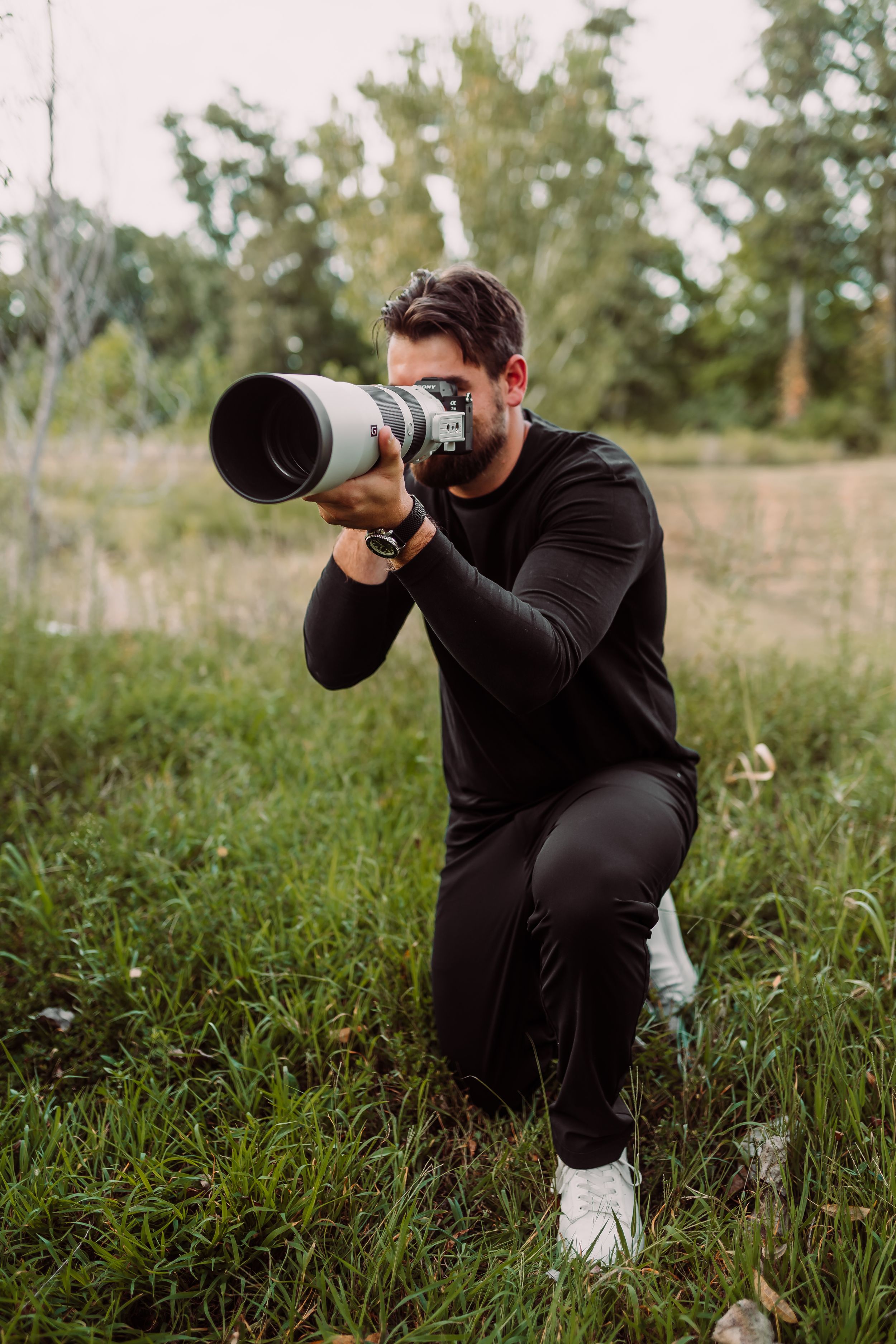 A photographer in black attire kneels in a grassy field while shooting with a large telephoto lens and camera.