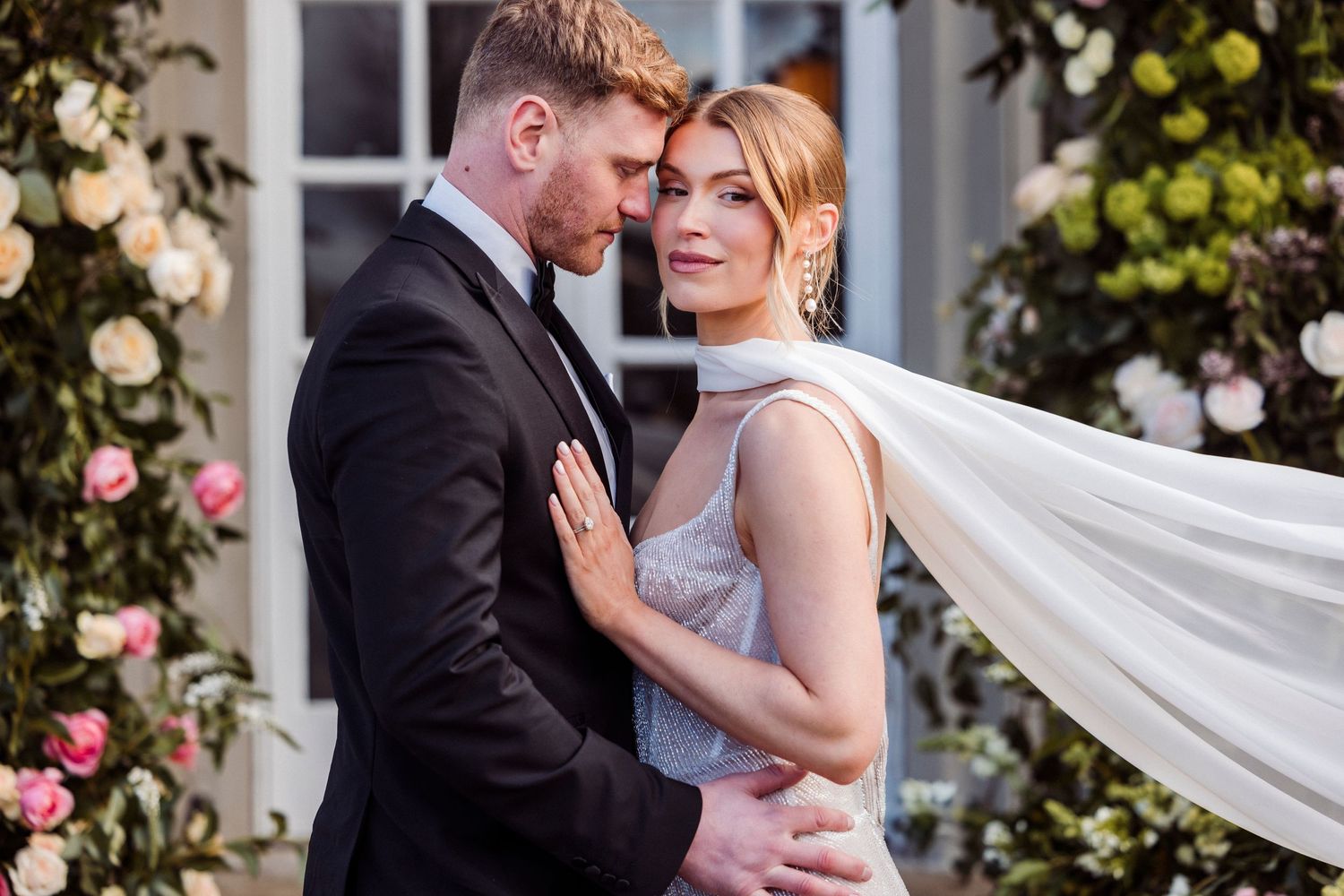 A romantic wedding couple embraces outdoors in front of Holesfoot adorned with climbing roses and greenery.