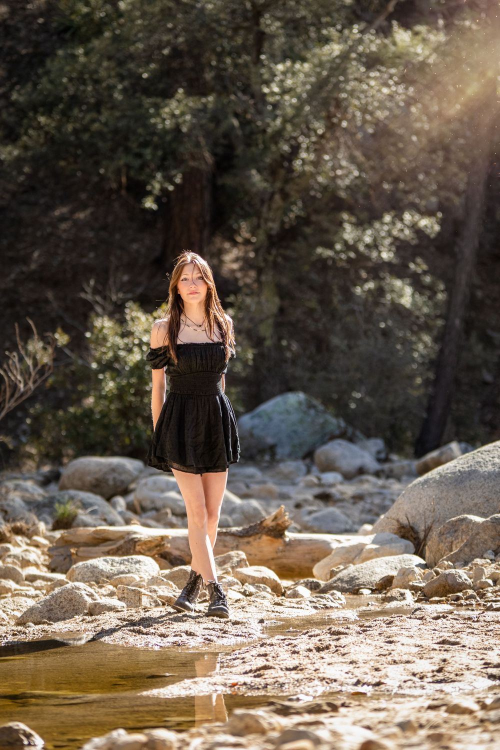 Person in black dress standing on rocks near flowing stream in sunlit forest setting.
