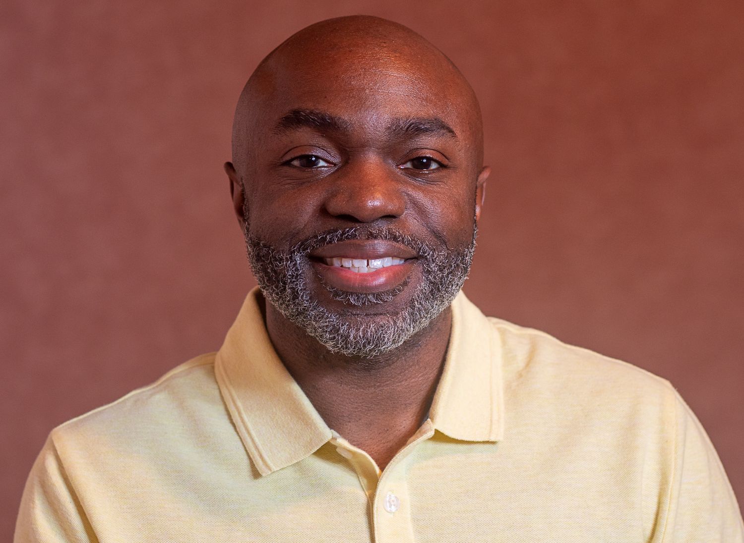 Man in yellow polo shirt smiling warmly against brown background.