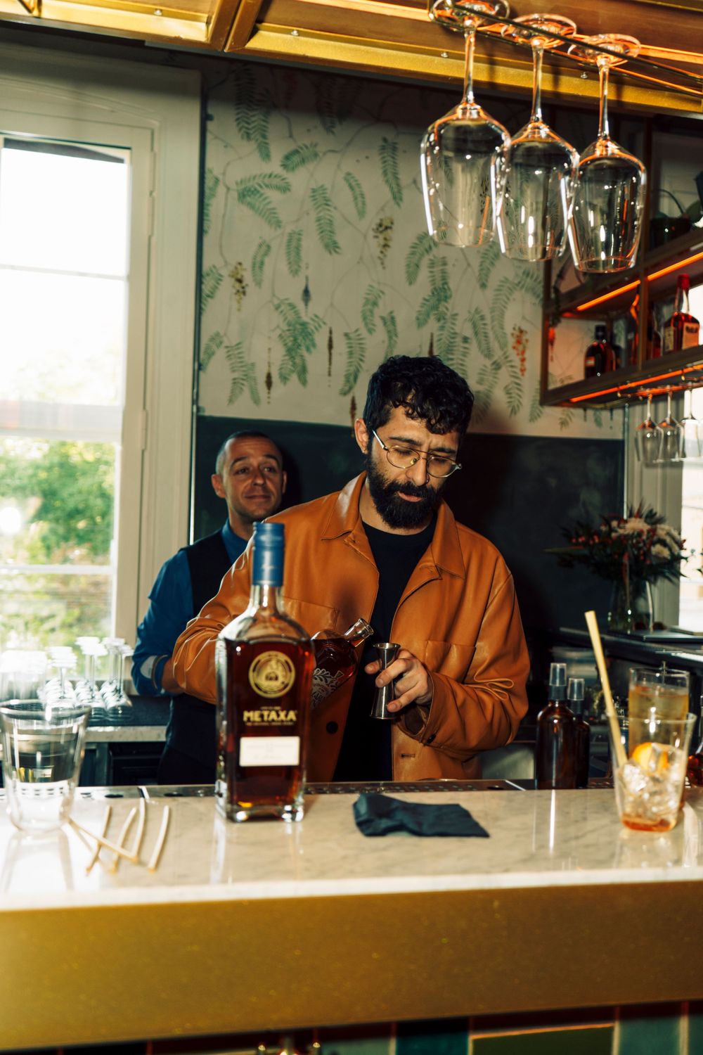 A cocktail maker behind a bar with a bottle of Metaxa on the countertop