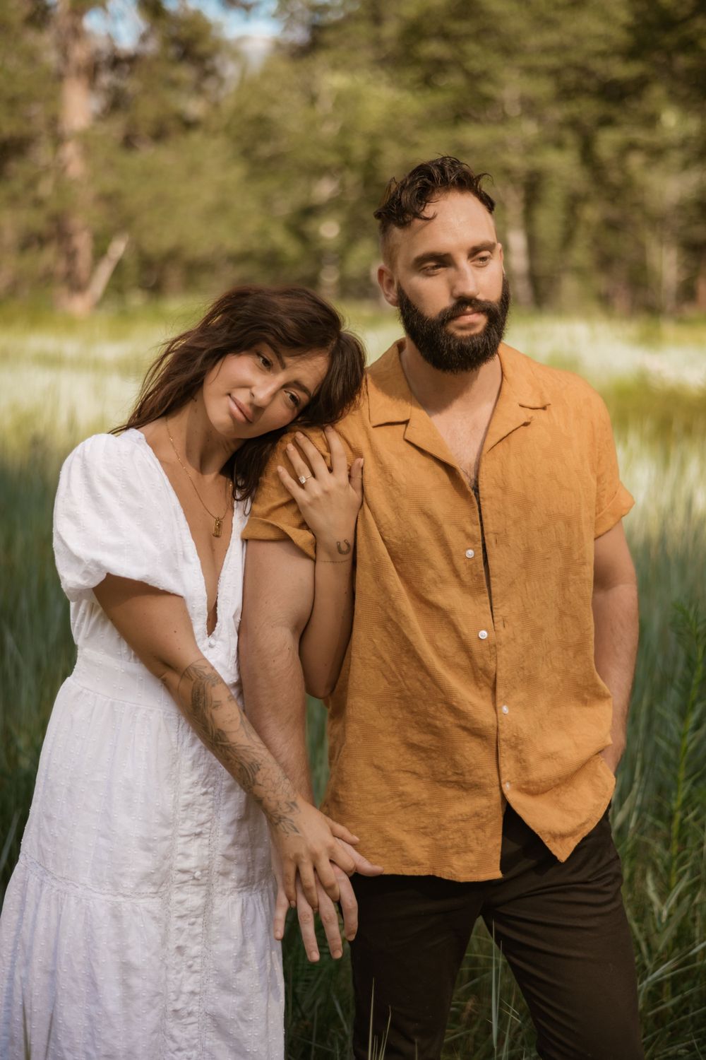 Intimate couple poses together in tall grass with golden light filtering through pine trees.