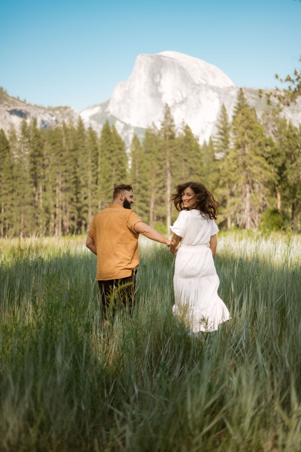 Couple walking through tall grass with snow-capped mountains and evergreen trees in the background.