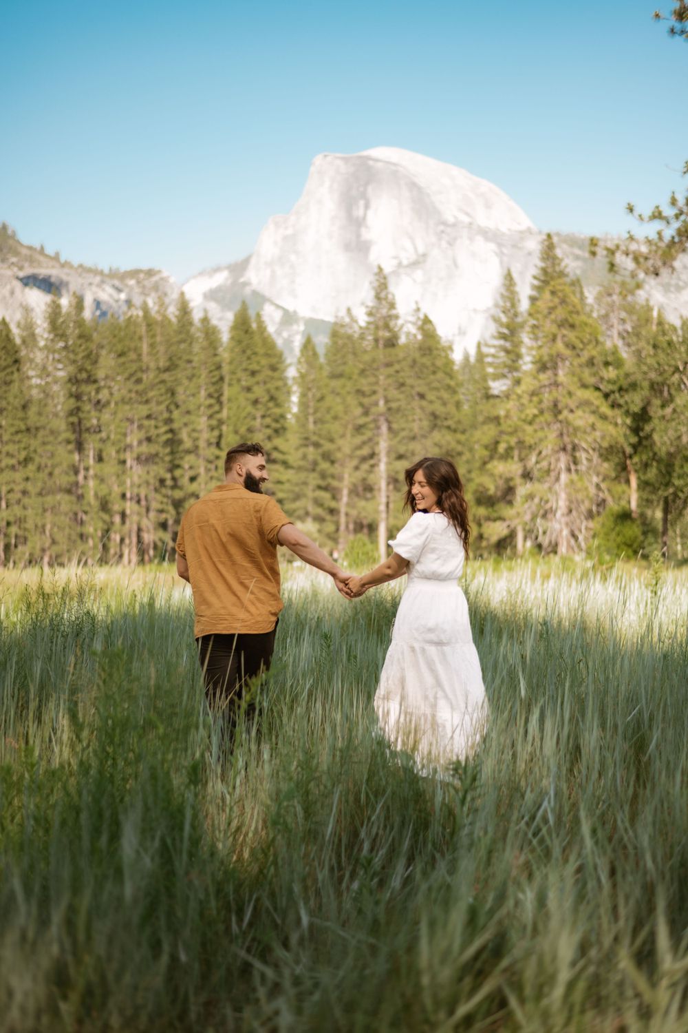 A couple dances playfully in a meadow with mountains and evergreen trees in the background.