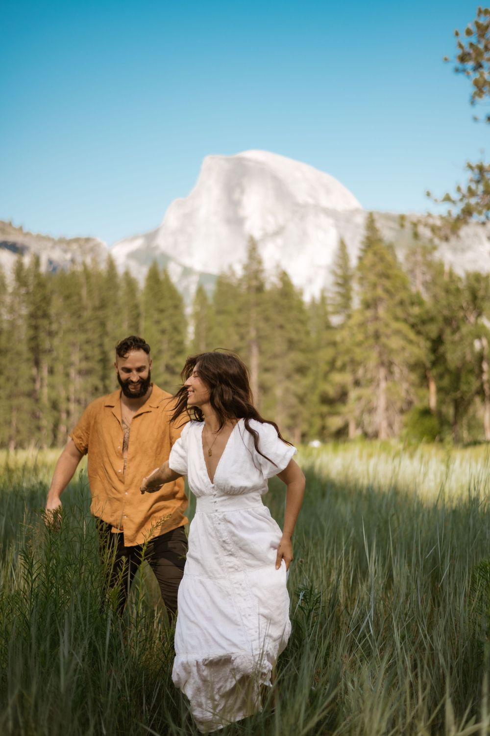 Couple runs playfully through tall grass with Half Dome visible in the mountain background.