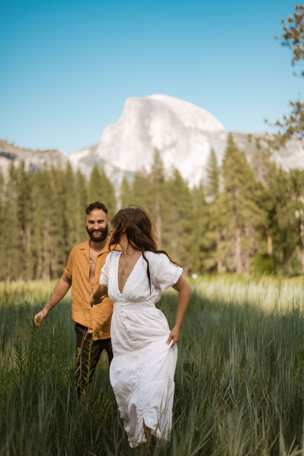 Two people run through tall grass with Half Dome mountain visible in Yosemite National Park.