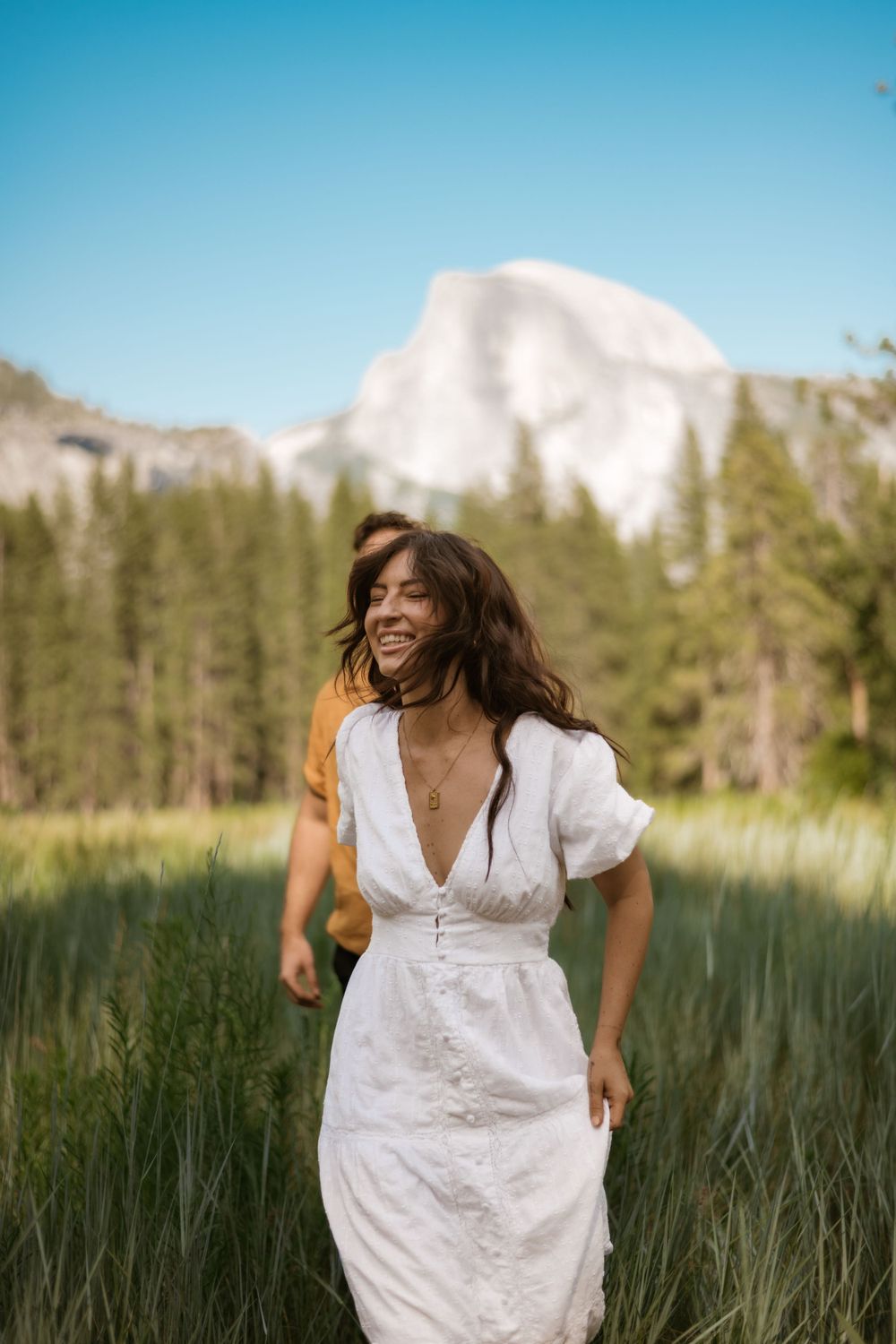 Woman in white dress twirling joyfully in meadow with Yosemite's Half Dome visible.