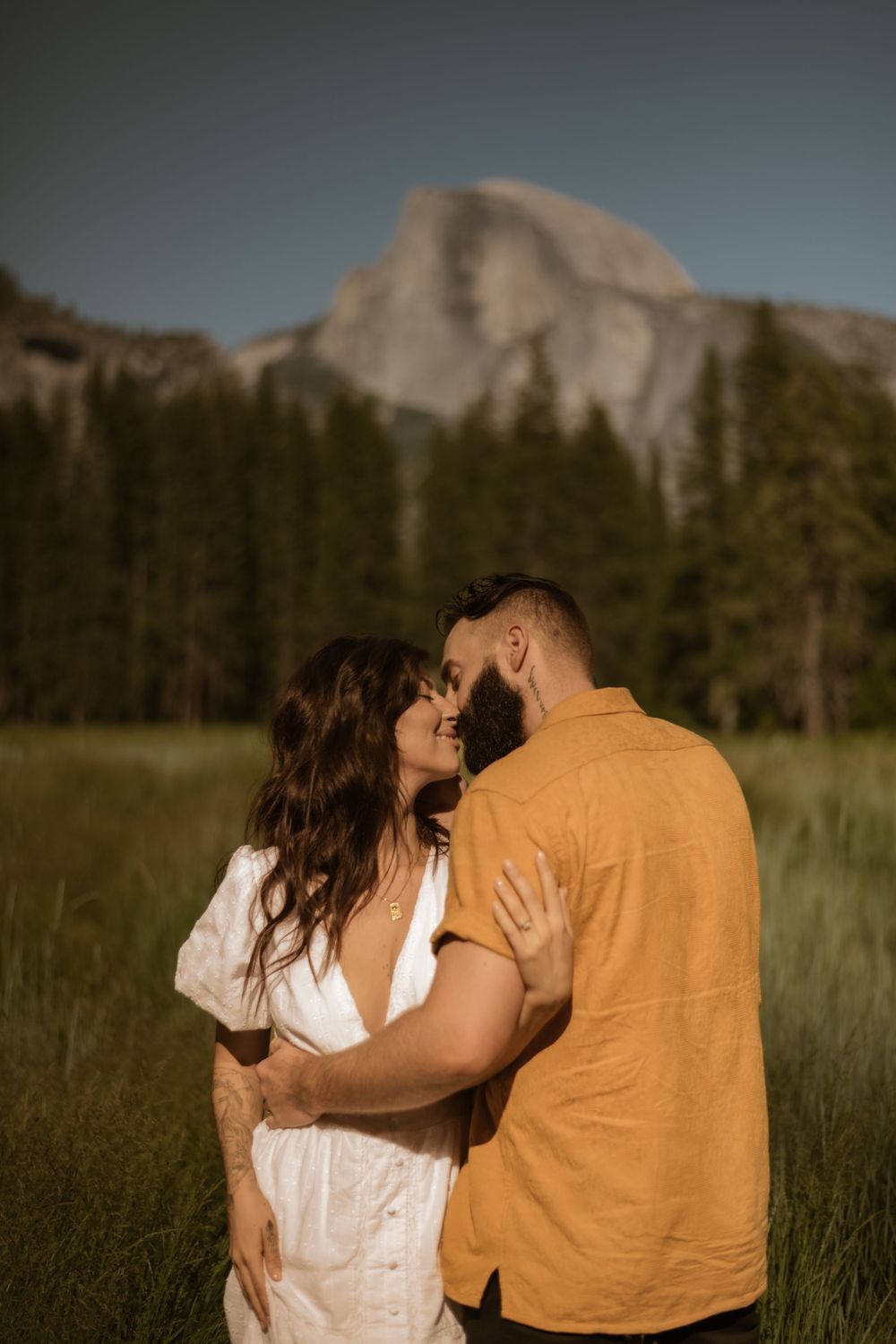 Couple sharing intimate moments in meadow with mountain peaks in background.