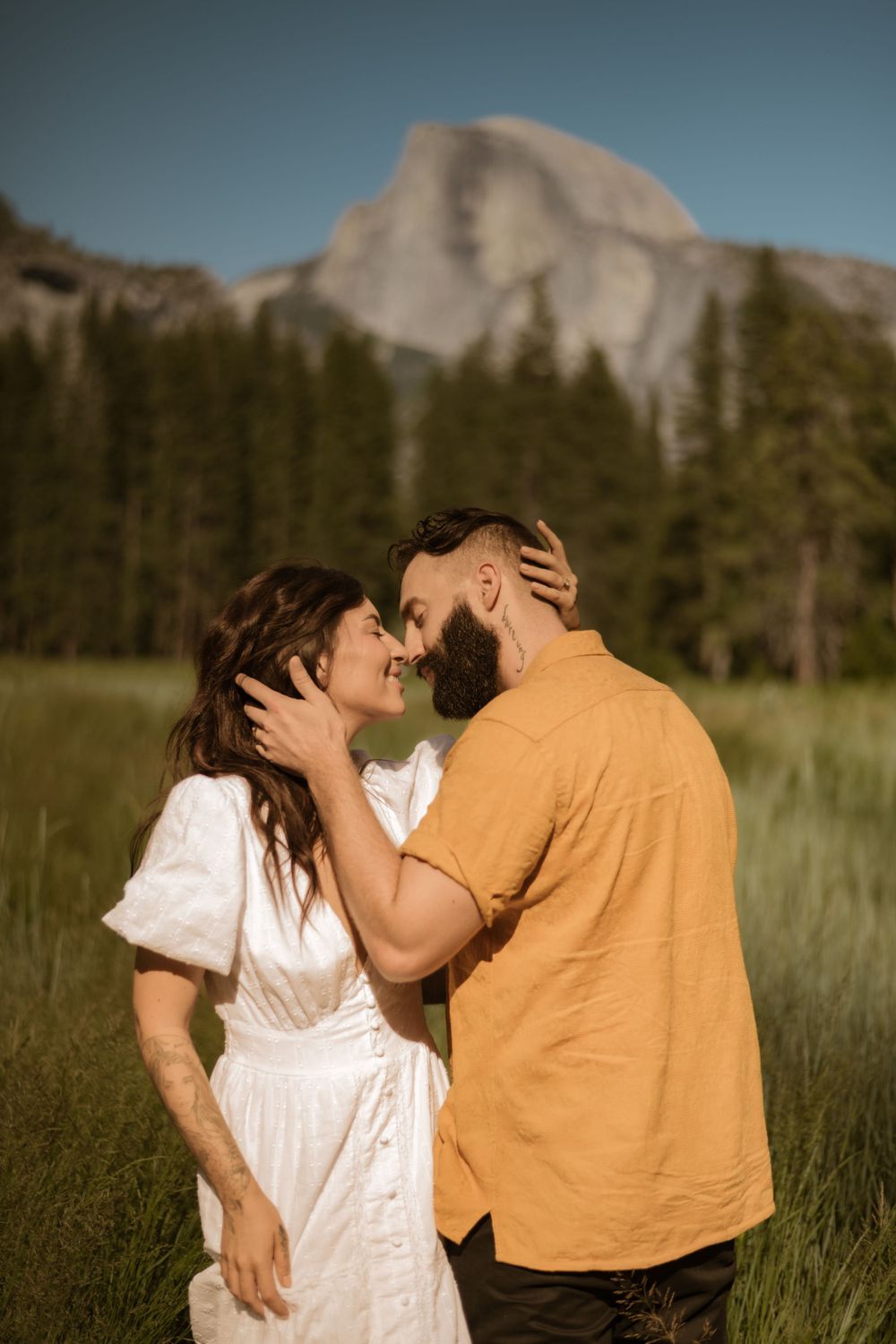 A romantic moment captured between two people in a sunlit meadow with mountains in the distance.