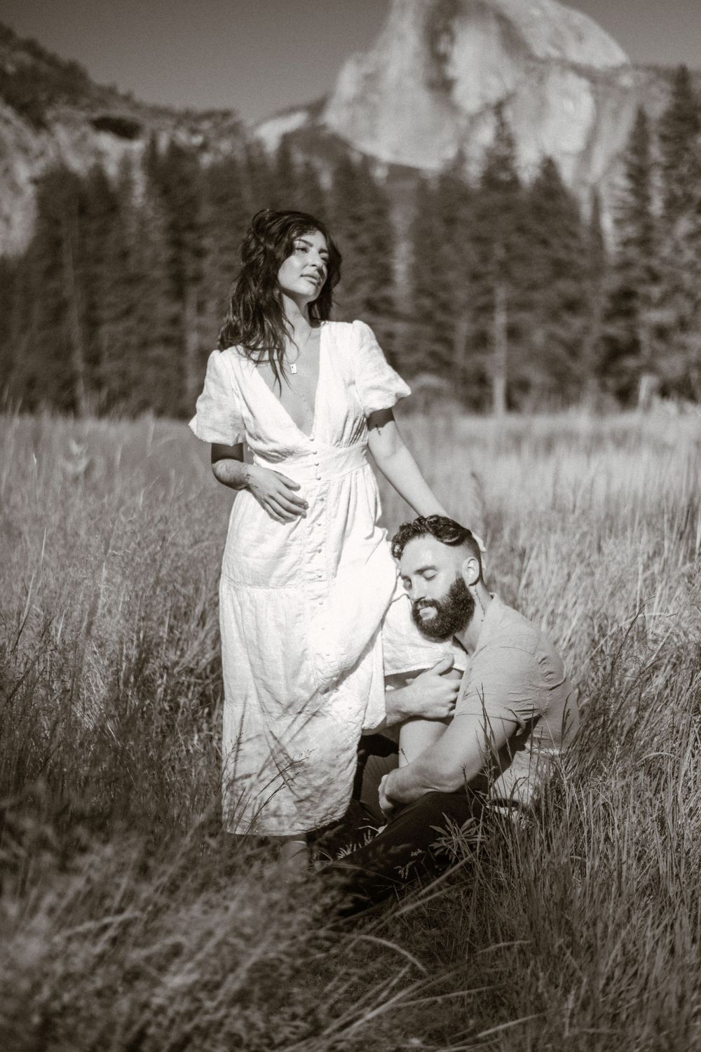 A couple shares an intimate dance in a grassy meadow with mountains and forest as their backdrop.