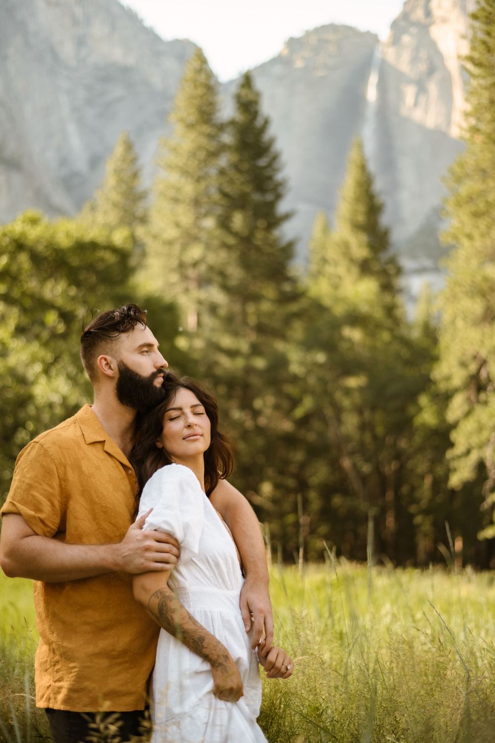 Sweet couple embraces in a grassy field with majestic mountain scenery behind them.