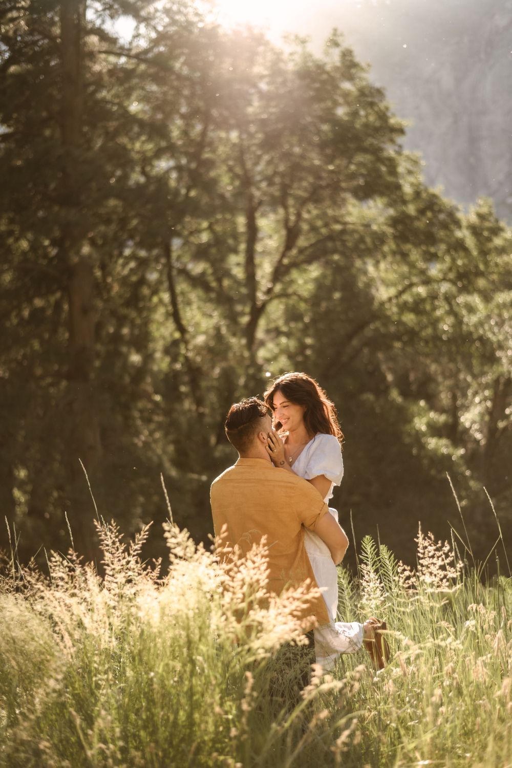 Romantic couple embraces in sun-drenched meadow with mountains and forest in background.