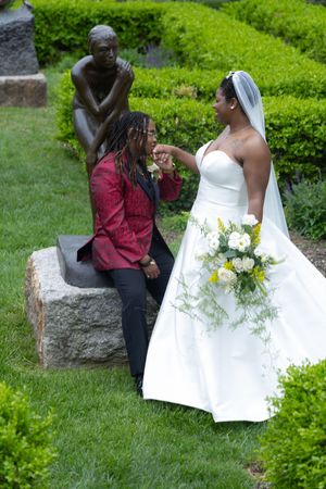 A bride in a white wedding dress holds a bouquet while someone kneels next to a garden statue.