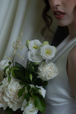 A delicate white bridal bouquet with roses and greenery held against a light colored dress.
