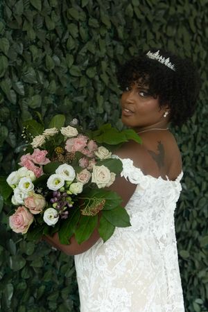 A bride in an off-shoulder lace gown poses with a pink and white floral bouquet against a leafy backdrop.