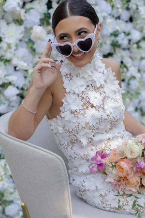 Bride in elegant white lace dress wearing heart-shaped sunglasses poses with bouquet against floral backdrop.