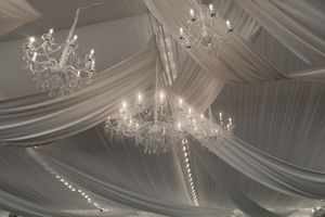 Elegant crystal chandeliers hang from a white draped ceiling at a wedding reception.