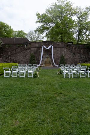 An outdoor wedding ceremony setup with white chairs and fabric arch against a stone wall.