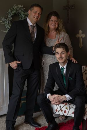 A formal portrait of three people posing together against a wall with decorative crosses in elegant evening attire.