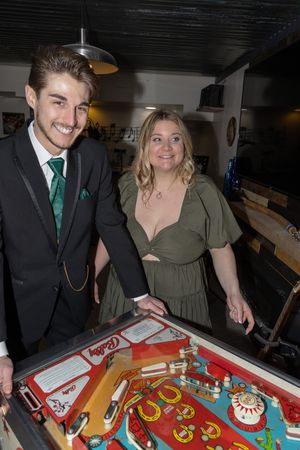 Two people smiling while playing pinball at an arcade bar with vintage machines in dim lighting.