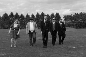 A black and white photograph shows five people walking together across a grassy field with trees in the background.