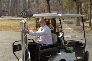 Person driving a golf cart along a paved path through a wooded area on a sunny day.