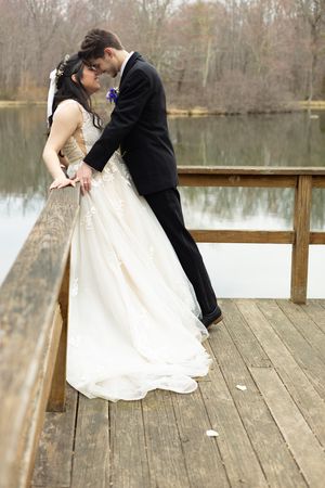 A couple in wedding attire embraces romantically on a wooden dock overlooking a serene lake during autumn.