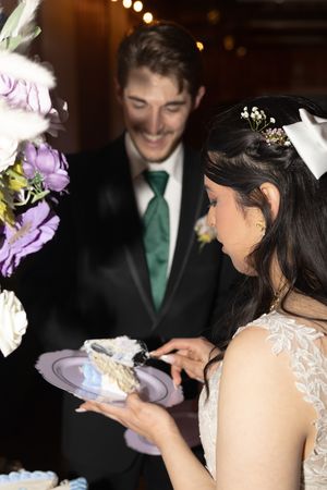 A couple shares a joyful moment while serving wedding cake at their reception, surrounded by purple floral decorations.