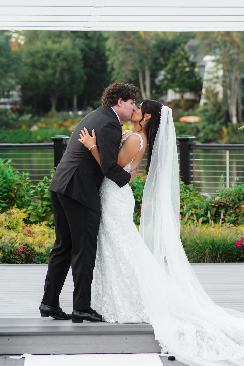 A romantic wedding moment captured on an outdoor deck with greenery and gardens in the background.