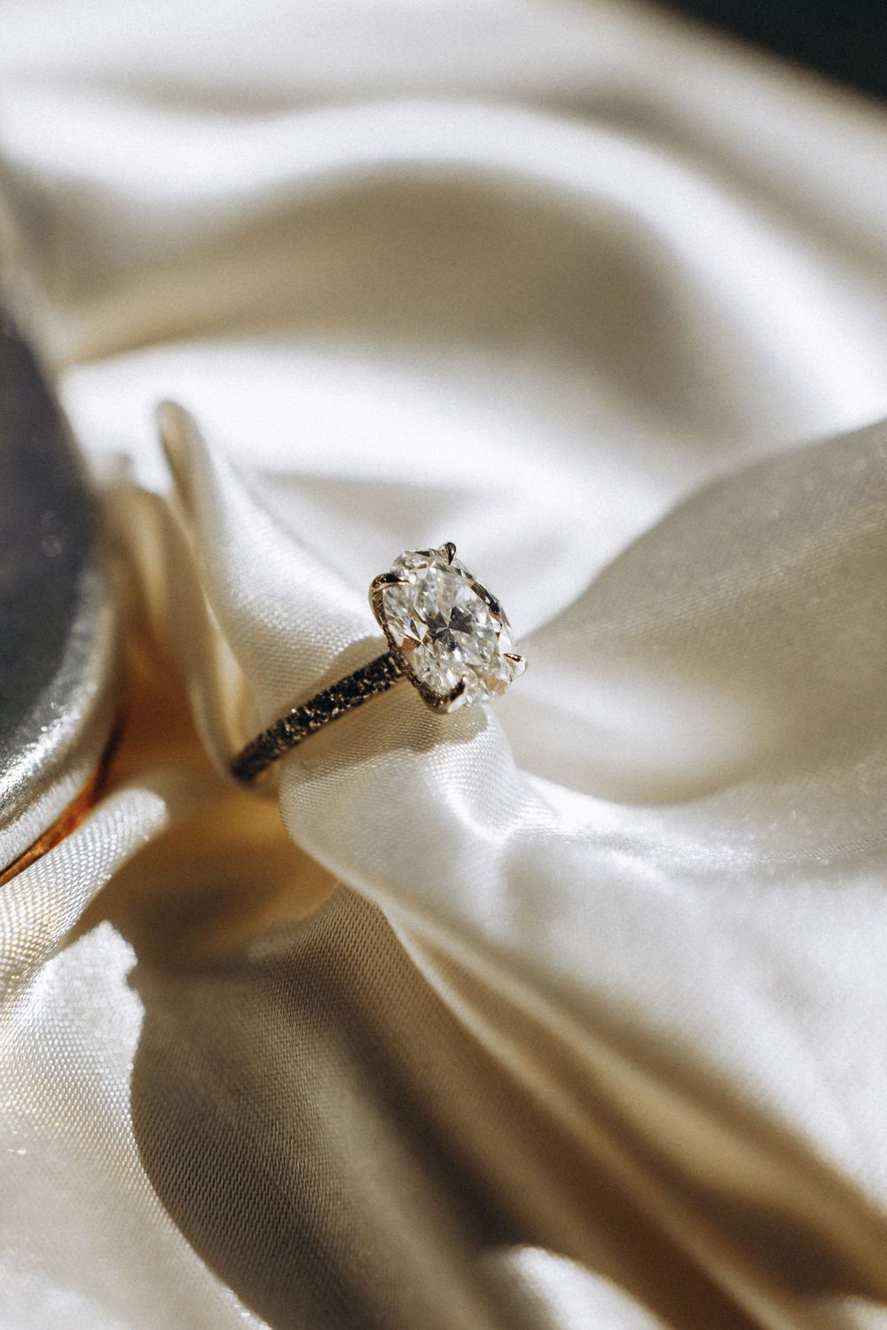 Elegant diamond engagement ring with oval cut stone rests on smooth white satin fabric in soft lighting.