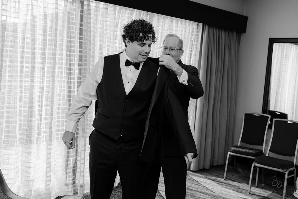 Two men in formal attire stand together in a black and white photo, one adjusting his tie while the other looks on.