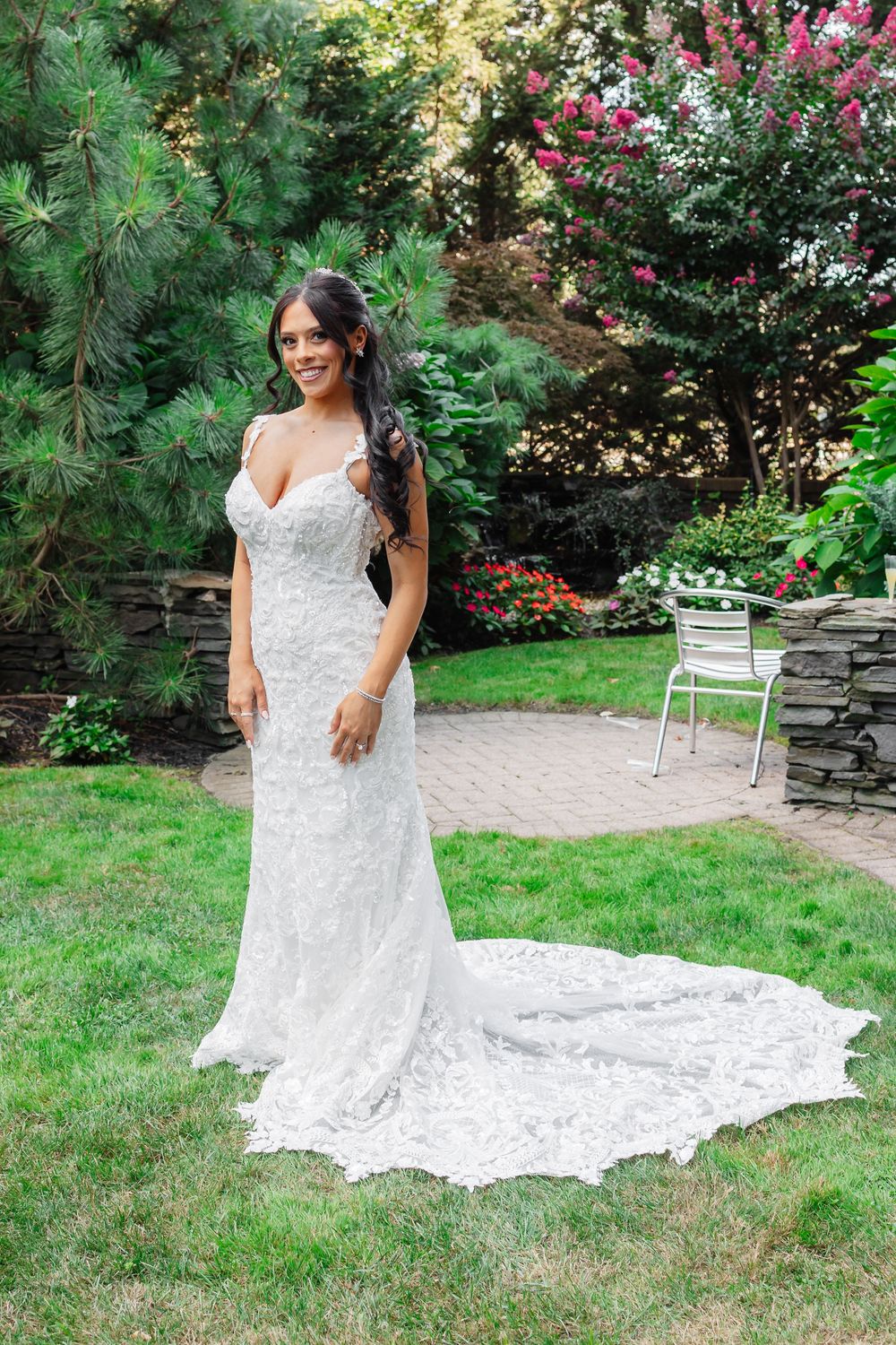 A stunning white lace wedding dress with a long train spreads across a garden lawn with flowering bushes in the background.