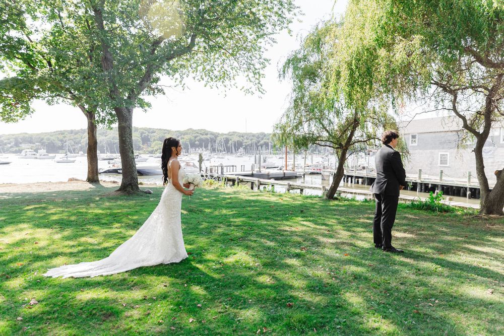 A couple in wedding attire poses for romantic photos on a grassy lawn with water and trees in the background.