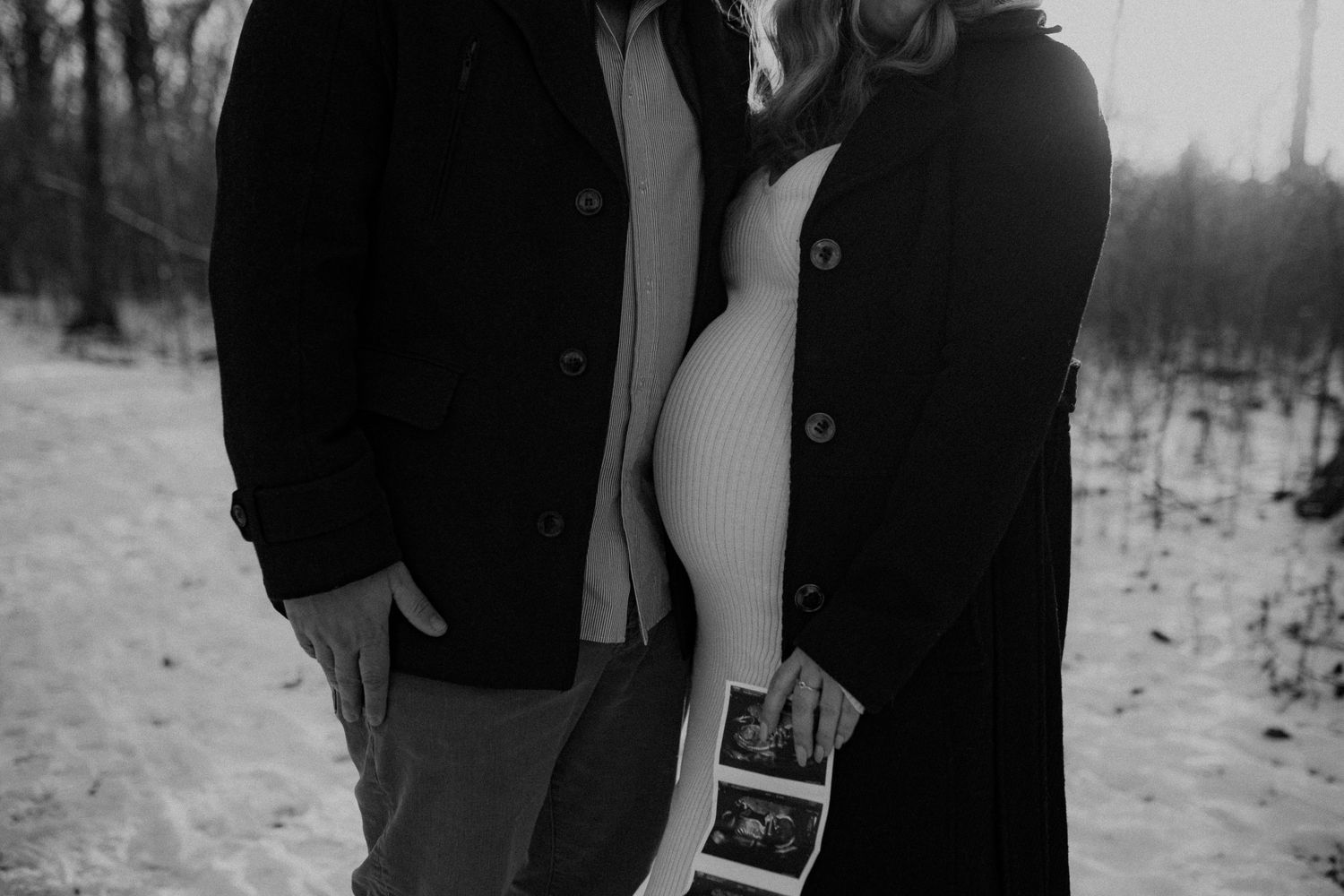 Couple standing in snowy winter forest holding ultrasound photos near exposed pregnant belly in black and white.