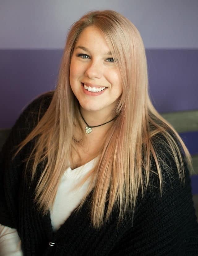 Portrait with long blonde hair wearing black cardigan against purple wall.