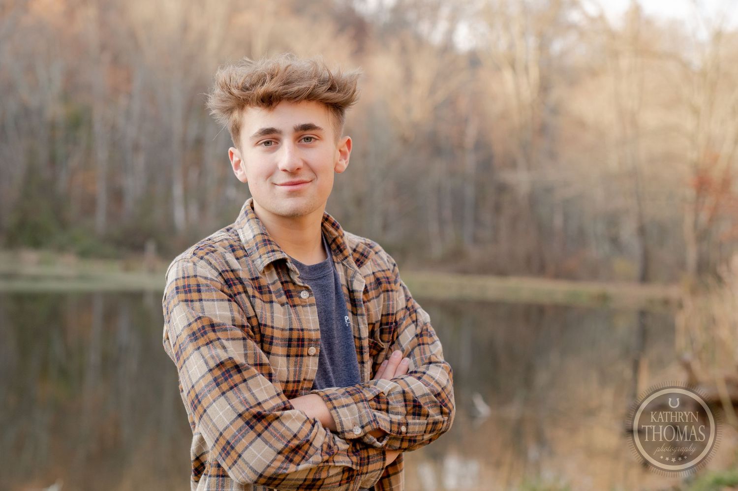 A person in plaid stands confidently with crossed arms near an autumn lake.