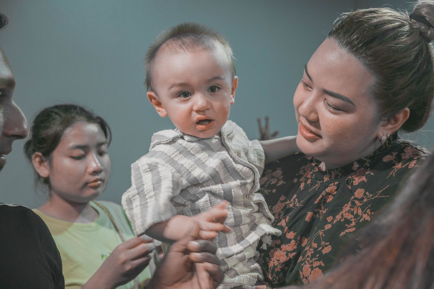 Small baby in white clothing being held during a family gathering.