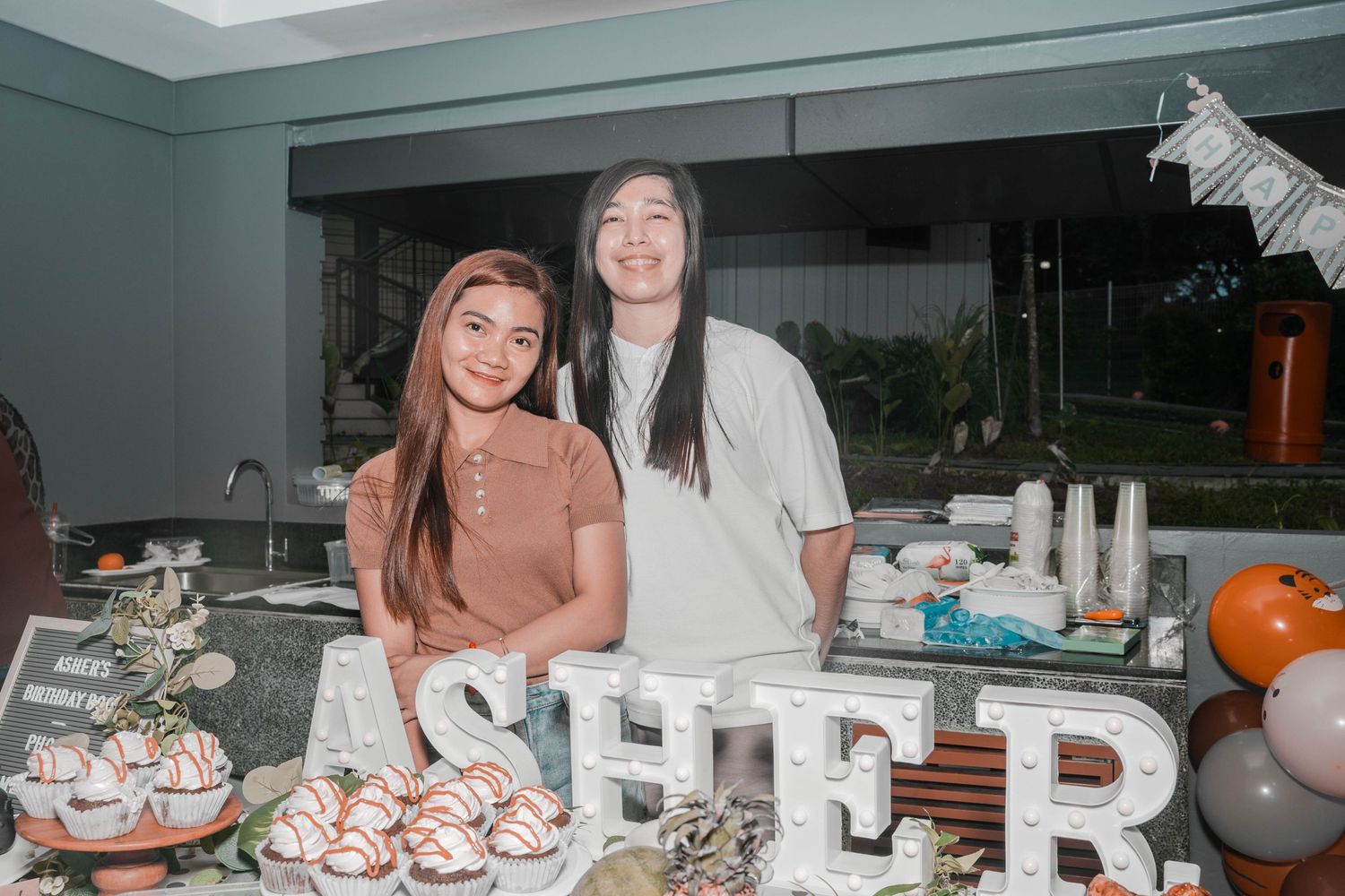 Two friends pose together at a party with decorative letters and balloons in the background.