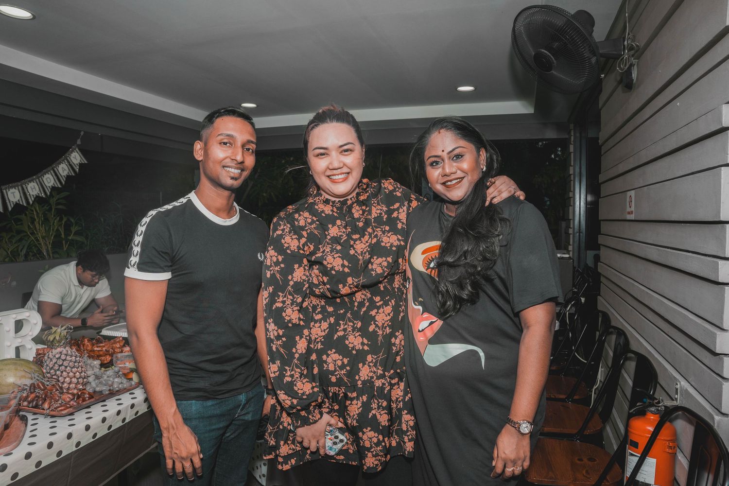 Three guests smile together at an evening celebration with party decorations.