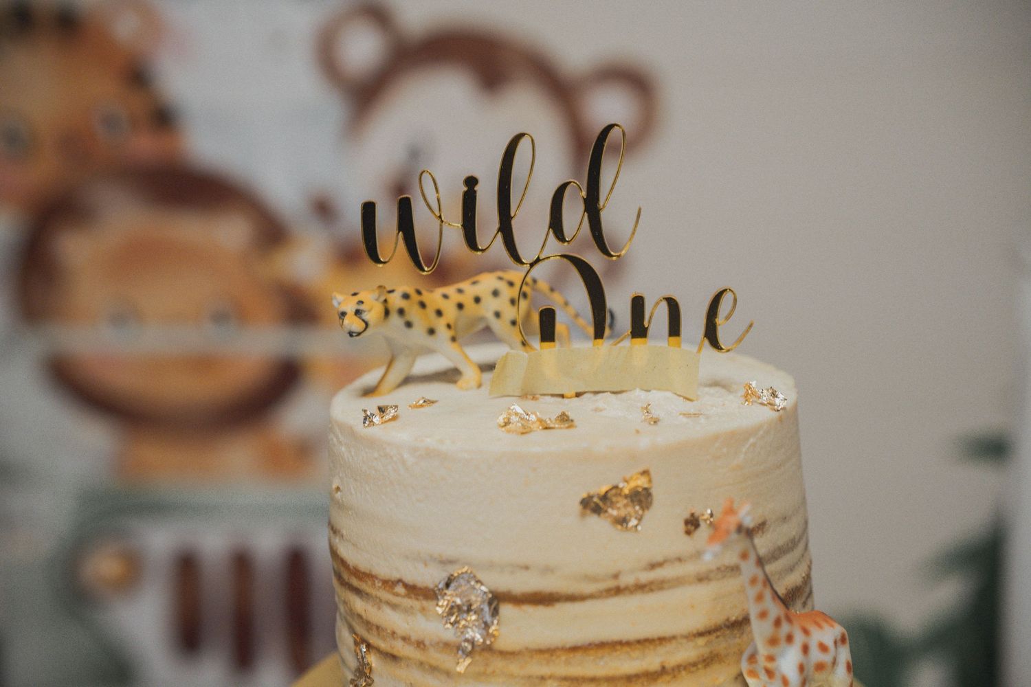 A close-up of a white birthday cake with a gold 'Wild One' topper and safari animal decorations.