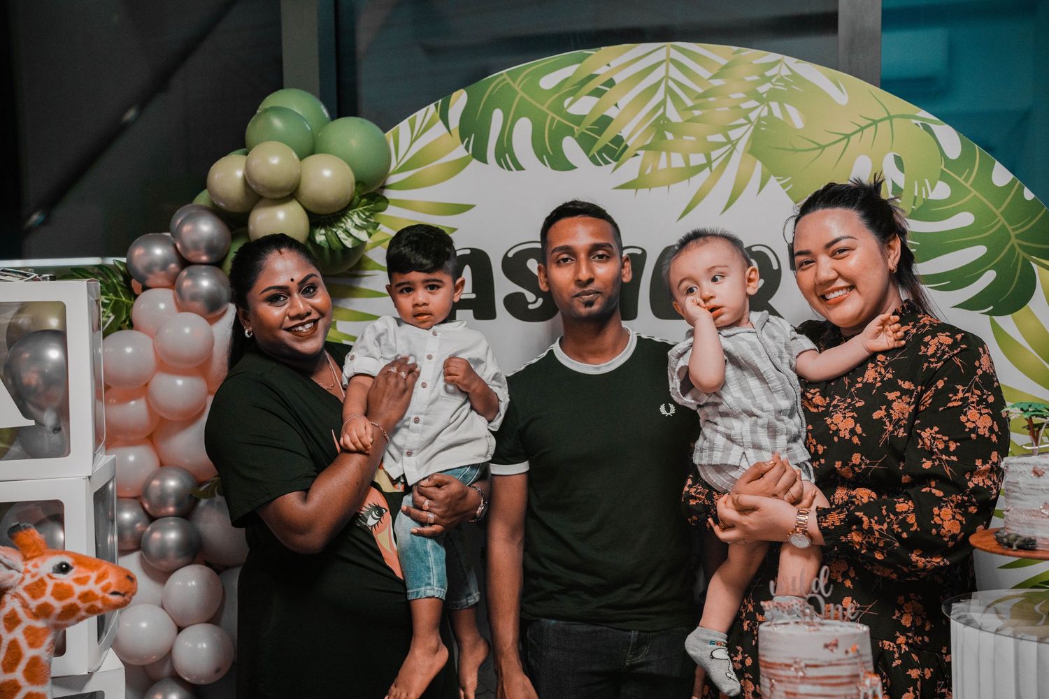 A group photo at a birthday party with tropical leaf backdrop and balloon decorations.