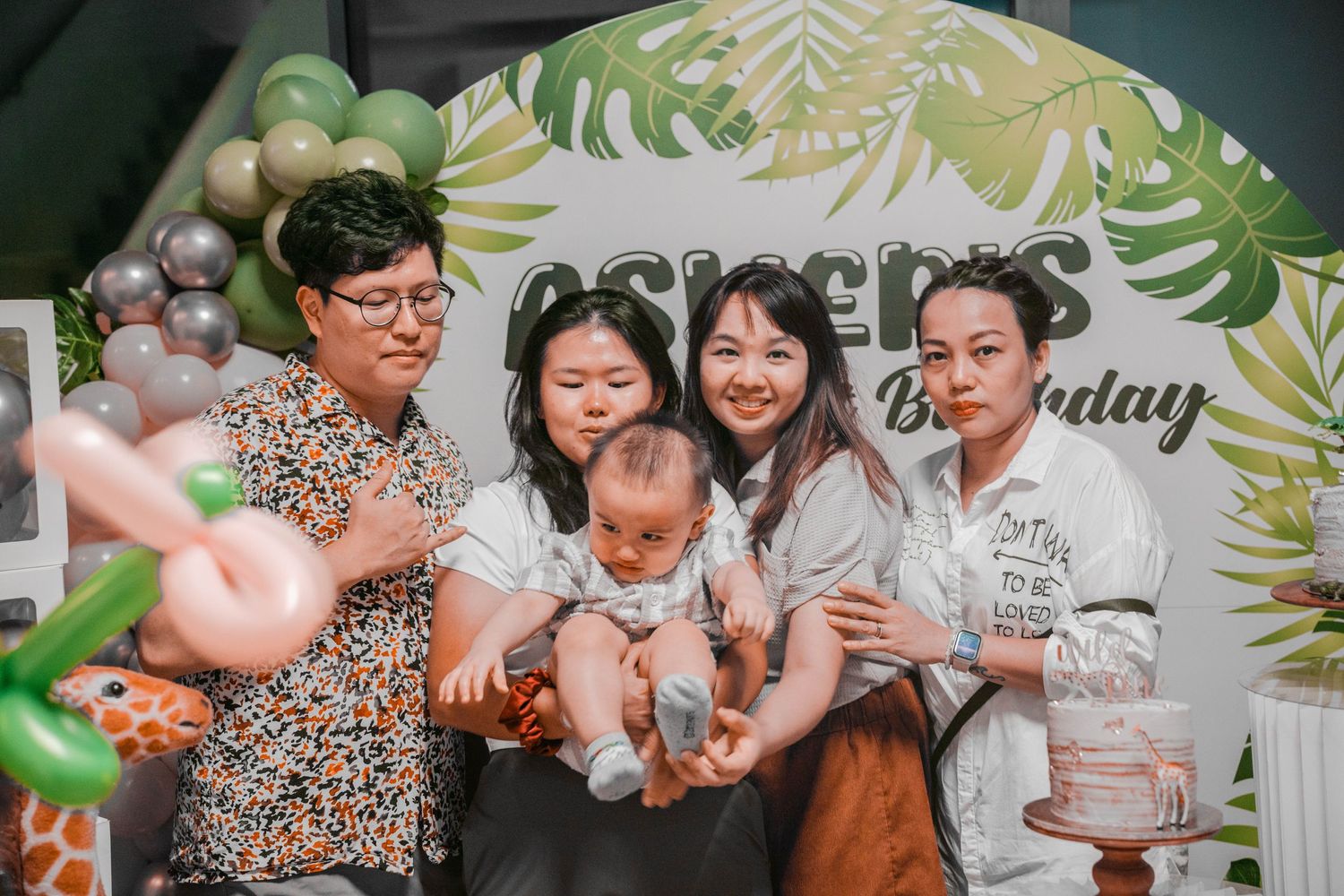 A group gathered around a baby at a birthday celebration with tropical themed decorations.
