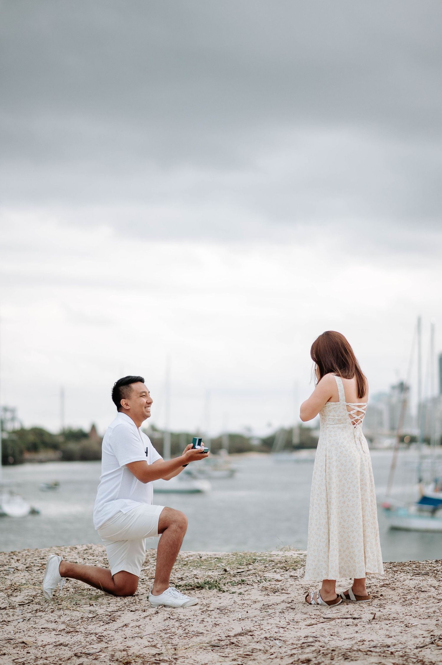 Couple getting engaged on rocky beach with cityscape and cloudy sky.